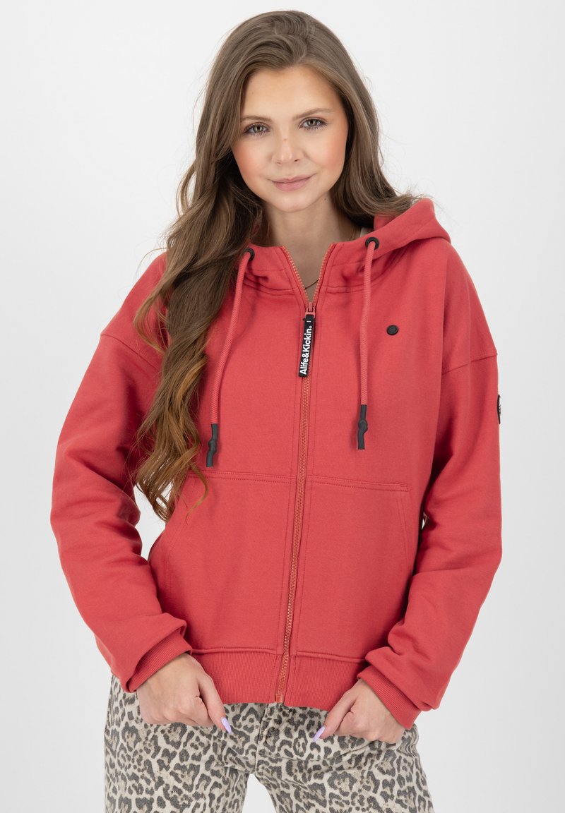 Felpa rossa con zip, realizzata in un tessuto morbido, con cappuccio regolabile, tasca frontale e dettagli neri. Abbinata a pantaloni con stampa leopardata.