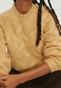 Pull en maille épaisse de couleur jaune doux, avec un motif en relief, un col côtelé et des manches surdimensionnées. Le matériau semble chaud et texturé.