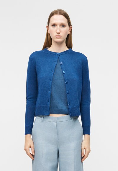 Femme aux cheveux roux raides portant un cardigan bleu boutonné sur un haut bleu et un pantalon gris clair, debout devant un fond uni.