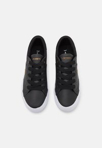 Zapatillas de cuero negro con suelas de goma, que presentan cordones negros, el logo dorado de Lacoste y una textura suave, diseñadas con una silueta de corte bajo.
