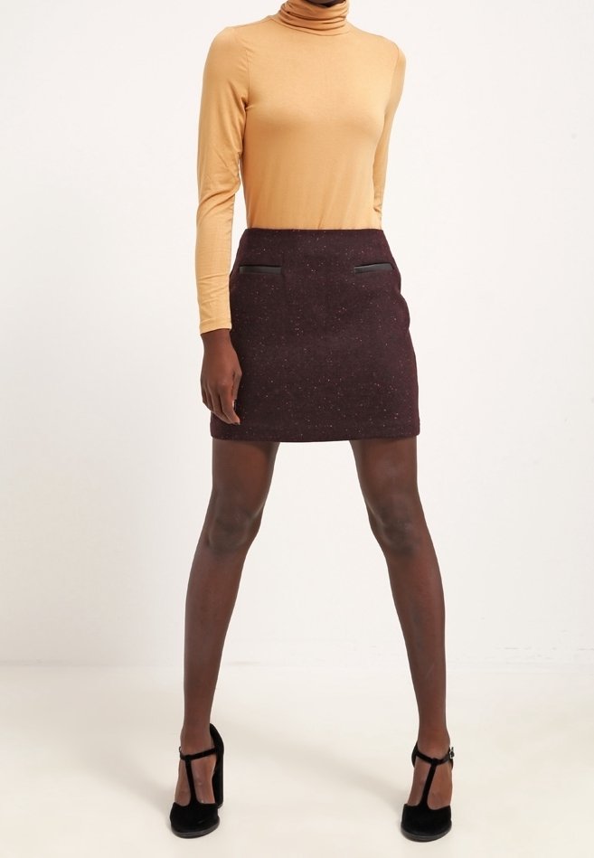 Femme portant un col roulé beige, une mini-jupe foncée tachetée avec poches avant, et des chaussures Mary Jane noires à talons hauts, debout jambes écartées.