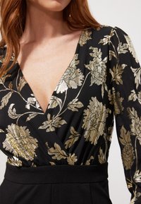 Top envolvente negro con estampado floral metálico dorado, escote en V profundo, mangas largas y cinturilla ajustada negra.