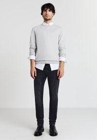 Grauer Crewneck-Pullover über einem weißen Hemd mit Kragen, kombiniert mit dunkler Jeans und schwarzen Stiefeletten, in einer minimalistischen Umgebung.