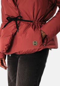 Rød isolert jakke med innsvingt midje, svart snor og logodetalj. Polstret tekstur og glatt finish. Kombineres med mørke jeans.