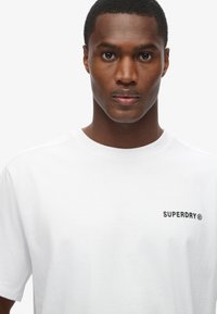 Hvid bomulds t-shirt med korte ærmer, der har et lille sort "SUPERDRY" logo på venstre brystområde. Glat tekstur, klassisk pasform.