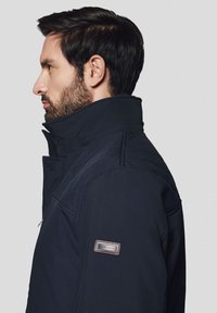 Granatowa kurtka ocieplana z wysokim kołnierzem, boczną kieszenią na zamek i pikowanymi detalami na ramionach. Naszywka z logo na lewym rękawie.