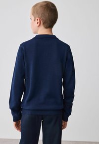 Granatowy sweter z dzianiny, z prążkowanym kołnierzem i mankietami. Posiada gładką fakturę oraz dopasowany krój z prostym dołem.