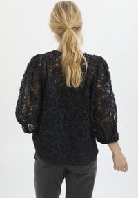 Svart blommig spetsblus med trekvarts puffärmar, texturerad yta och genomskinlig rygg. Matchad med grå jeans, sedd bakifrån.