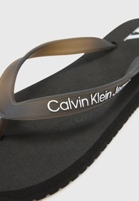 Juodos šlepetės su matinio paviršiaus tekstūra, turinčios permatomą rudą dirželį ir baltą "Calvin Klein Jeans" logotipą ant diržo.