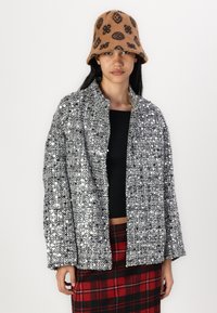 Veste ornée de sequins en noir et argent, présentant une coupe ample et un design à l'avant ouvert. Associée à un haut noir et une jupe à carreaux.