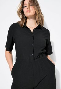Mono corto negro de manga corta con cuello tipo polo, frente con botones, cintura elástica y bolsillos laterales; tejido texturizado.