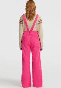 Person med langt rødt hår har på seg en beige langermet skjorte med stripete ermer og lyse rosa vide bukser med seler, og står med ryggen til.