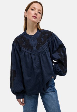 Blouse bleu marine avec un col rond, des manches bouffantes et une broderie florale noire sur les épaules. Elle comporte une fermeture par boutons et une texture à pois.