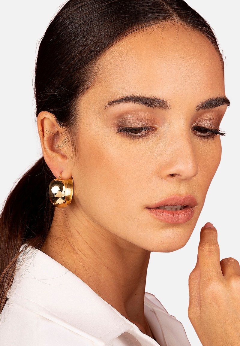 Des boucles d'oreilles en or avec une surface brillante et texturée, présentant un design arrondi. Le modèle a de longs cheveux et un maquillage neutre.