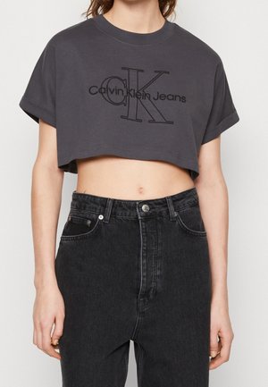 Kobieta ubrana w ciemnoszarą krótka koszulkę Calvin Klein Jeans i czarne dżinsy z wysokim stanem, stojąca z ramionami luźno opuszczonymi wzdłuż ciała.