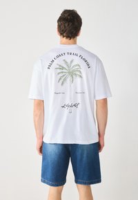 Valge puuvillane t-särk, mille seljal on palmipuude graafika, peal on tekst "PALM COAST TRAIL FLORIDA" ja all "Key West" allkiri.