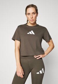 Majica s kratkim rukavima, skraćena, atletska u tamnosmeđem pamuku s bijelim geometrijskim logotipom sprijeda. Uparena s odgovarajućim maslinasto zelenim tajicama.