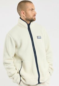 Fleecejacke in Off-White mit hohem Kragen, durchgehendem Reißverschluss und navyfarbenen Akzenten. Verfügt über seitliche Taschen und ein Berglogo auf der Brust.