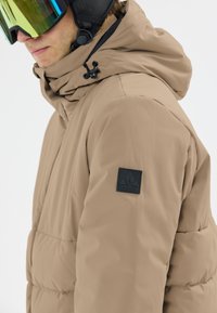 Beige Steppjacke mit hohem Kragen und verstellbare Kapuze. Verfügt über ein schwarzes Patch-Logo auf dem linken Ärmel und eine glatte Außenstruktur.