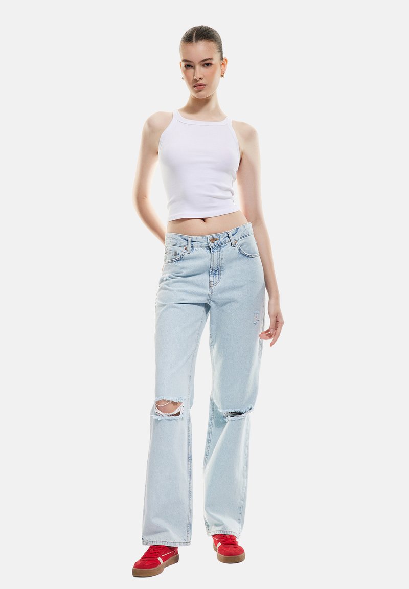 Top corto blanco, jeans azul claro desgastados con rasguños en las rodillas y zapatillas rojas con suelas marrones. Diseño simple y moderno.