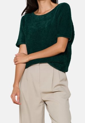 Zielony, futrzasty sweter o krótkich rękawach i szerokim dekolcie, w zestawie z jasnobeżowymi, wysokim stanem spodniami. Gładka faktura, luźny krój.