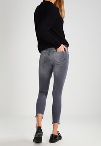 Jersey negro ajustado, jeans de mezclilla gris con un dobladillo desgastado y zapatos planos negros. Los jeans tienen dos bolsillos traseros y un corte ajustado.