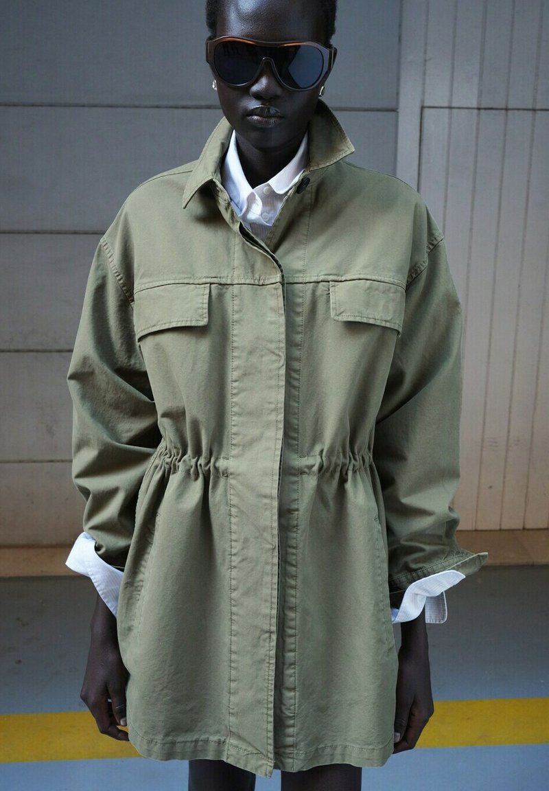 Dunkelhäutige Person mit großen schwarzen Sonnenbrillen, weißem Hemd und übergroßer hellolivgrüner Jacke mit elastischem Bund, steht drinnen.