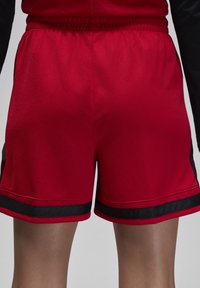 Punaiset urheilushortsit, jotka on valmistettu teksturoidusta kankaasta. Niissä on mustat sivukoristeet ja musta joustava vyötärönauha, keskireisien pituinen malli.