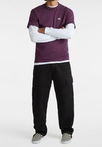 Fialové tričko s krátkým rukávem vrstvené přes bílé tričko s dlouhým rukávem, v kombinaci s černými cargo kalhotami a černobílými kockovanými nazouvacími botami.