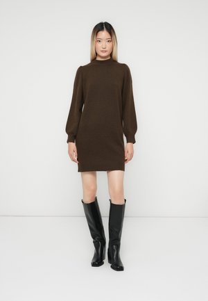 Frau, die vor einem weißen Hintergrund steht und ein braunes Strickkleid mit langen Ärmeln sowie schwarze, knielange Lederstiefel trägt.