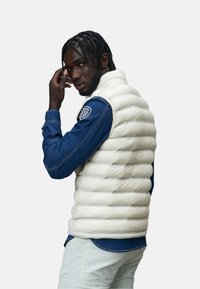 Gilet piumino bianco con colletto alto, caratterizzato da sezioni trapuntate, indossato sopra una camicia di jeans blu con cuciture a contrasto e un toppe decorative sulle maniche.