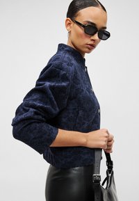 Σκούρο μπλε ναυτικό μπουφάν με υφή, μανδύας μανταρινιού και κλείσιμο με κουμπιά, σετ με λεπτό μαύρο δερμάτινο παντελόνι και μαύρη τσάντα.