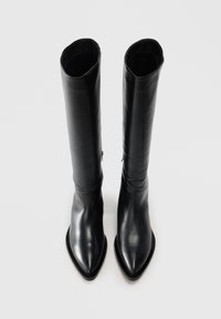Une paire de hautes bottes en cuir noir avec des pointes aiguës et des fermetures éclair sur les côtés, présentées debout sur un fond gris clair.