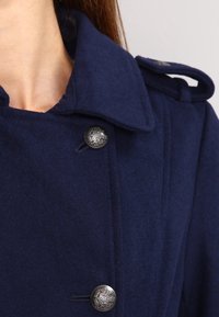 Marinblå ullkappa med stora svarta knappdetaljer, strukturerad krage och axelklaffar. Mjuk textur och skräddarsydd design.