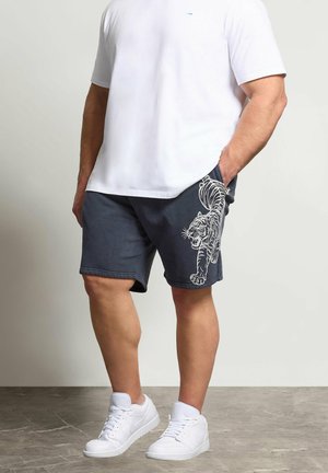 Homme portant un t-shirt blanc, un short bleu marine avec un motif de tigre blanc sur la jambe gauche, et des baskets blanches, debout avec une main dans la poche.