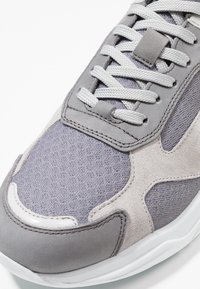 Baskets athlétiques grises avec un mélange de maille texturée et de daim lisse, accentuées de gris clair et dotées de lacets plats. Semelle en caoutchouc blanche visible.