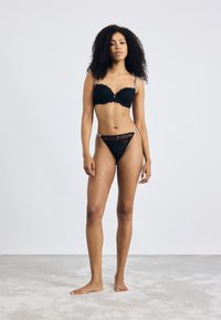 Black lace push-up bra with scalloped edges and silver accents, paired with matching black lace thong. Model standing on a light carpet.