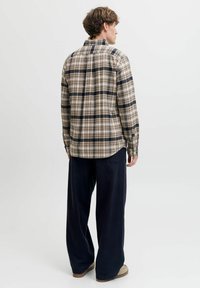 Młody mężczyzna stojący tyłem, ubrany w beżową i granatową koszulę w kratę, luźne ciemnogranatowe spodnie i beżowe buty na białym tle.