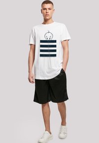 Hvit t-skjorte med et grafisk motiv av en karakter over horisontale marineblå striper. Modellen har på seg svarte shorts og hvite joggesko.