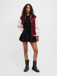 Bordo in krem varsity jakna s črtastimi robovi, črn prilega majica, razširjena črna krila in rjavi škornji do kolen. Podrobnosti vključujejo logo na jakni.