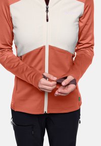 Giacca con zip arancione e crema caratterizzata da una parte superiore testurizzata, maniche aderenti e aperture per il pollice, abbinata a pantaloni scuri.