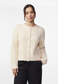 Cremfarbener strick cardigan mit einem strukturierten, diamantartigen Muster, gewelltem Ausschnitt und großen Perlenknöpfen auf der Vorderseite. Die Ärmellänge ist lang.