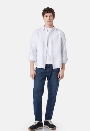 Młody mężczyzna w białej koszuli w paski zapinanej na guziki, na białej koszulce, niebieskich dżinsach i czarnych trampkach, stojący na jednolitym tle.