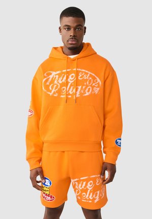 Mężczyzna w jaskrawopomarańczowej bluzie z kapturem i dopasowanych szortach z białymi i wielokolorowymi logo "True Religion", stojący na jednolitym tle.