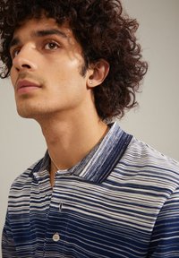 Polo rayé en bleu et blanc, avec un col, des boutons et un tissu texturé. Le modèle a des cheveux bouclés et une petite boucle d'oreille en forme de cœur.