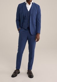 Granatowy garnitur trzyczęściowy z dopasowaną marynarką, kamizelką i wąskimi spodniami. Biała koszula pod spodem, zestawiona z brązowymi mokasynami. Gładka faktura materiału.