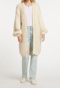 Chunky gebreid crèmevest met een losse pasvorm, geribbelde textuur en een open voorkant. Gecombineerd met lichtblauwe distressed jeans en witte sneakers.