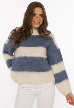 Frau mit langem braunem Haar, die einen blau-weiß gestreiften flauschigen Pullover und weiße Hose trägt, lächelt und hat eine Hand in der Hüfte.