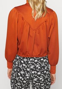 Blusa de color óxido con mangas abullonadas y un yugue trasero en forma de V, combinada con pantalones negros estampados con flores, que cuentan con acentos blancos.