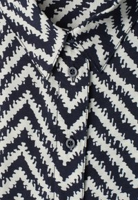 Col de chemise avec un motif zigzag noir et blanc et deux boutons noirs, montrant un tissu texturé et des détails de couture.